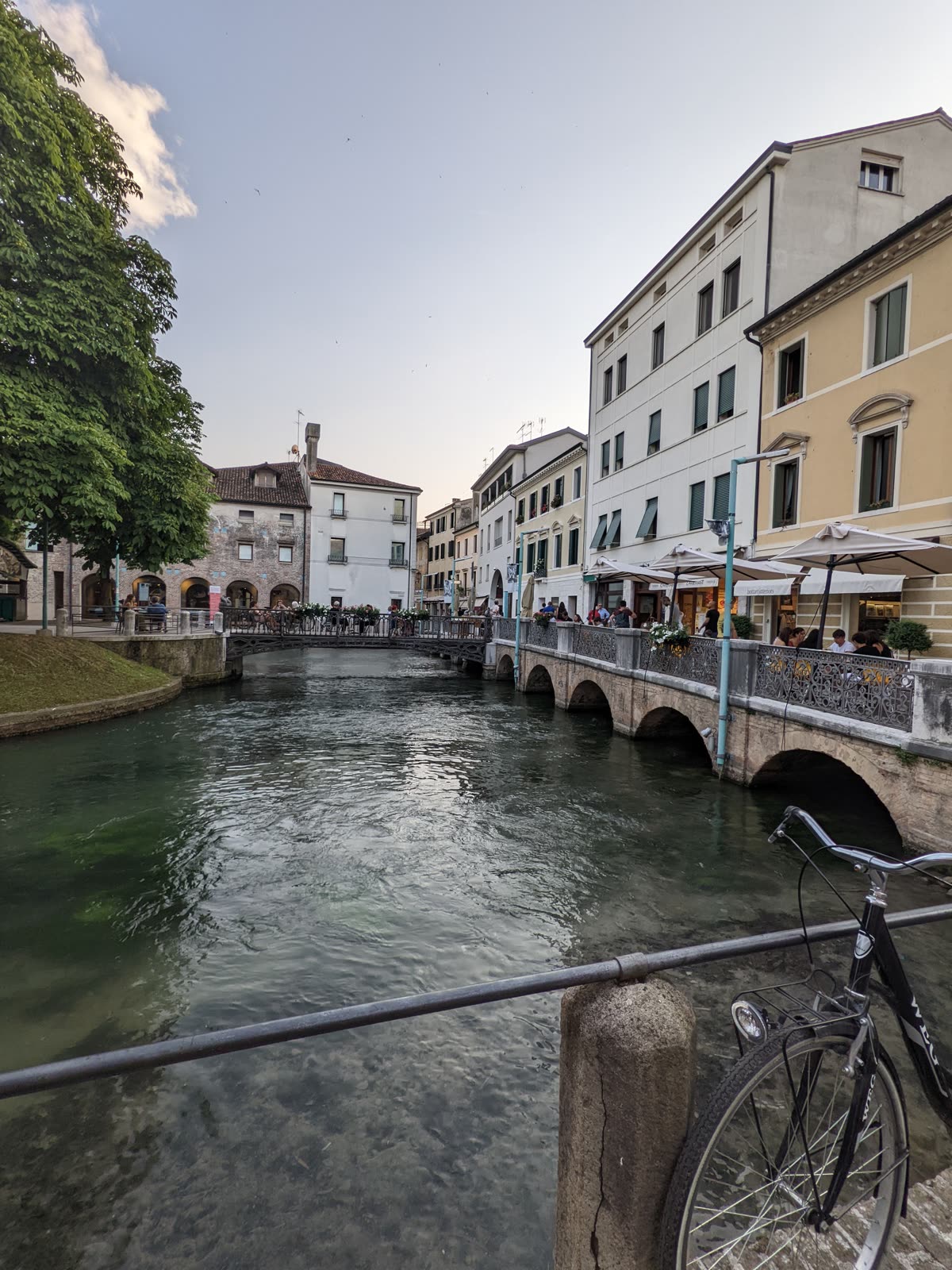 A canal in Treviso's old center at dusk, a low arched stone footbridge crossing it, café umbrellas and diners along an arcaded waterfront, historic pastel buildings, a bicycle leaning on the near railing