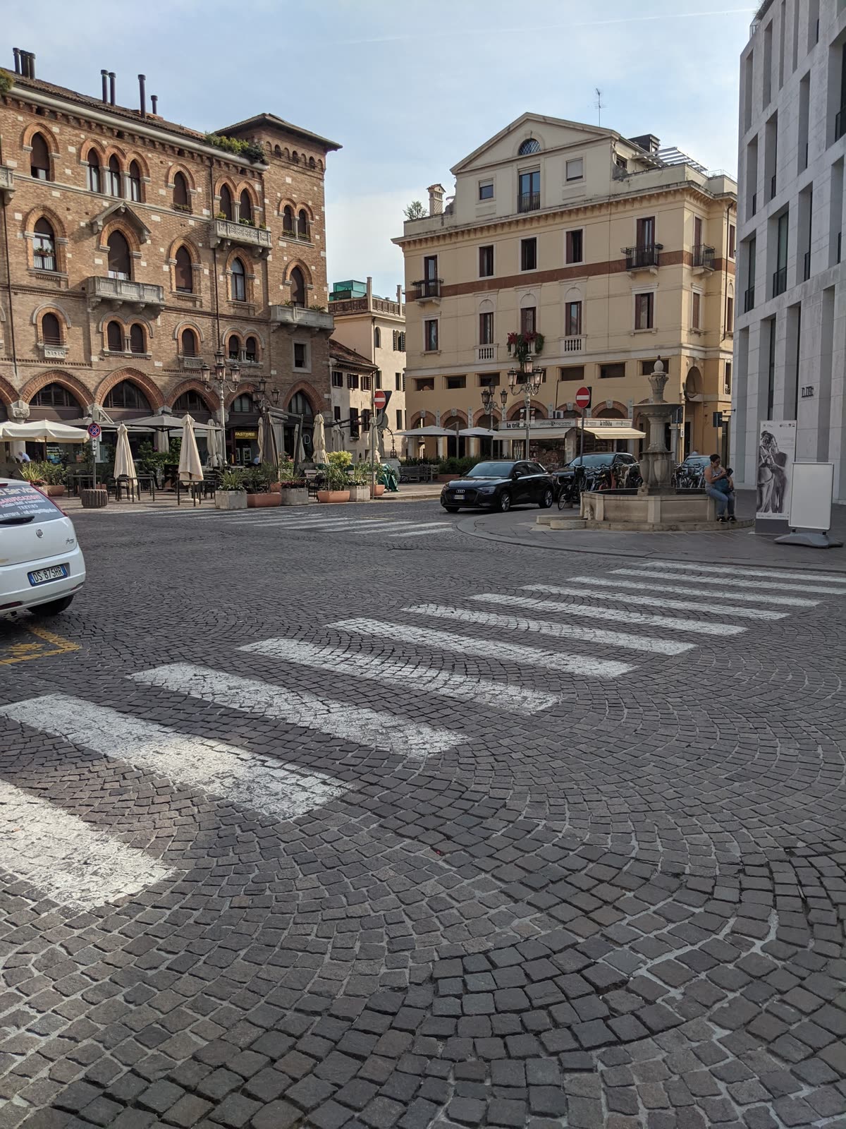 A cobbled Treviso piazza in early morning light, an old octagonal marble fountain at right, café umbrellas still folded under the arcades of historic brick and stucco buildings, a couple of cars crossing