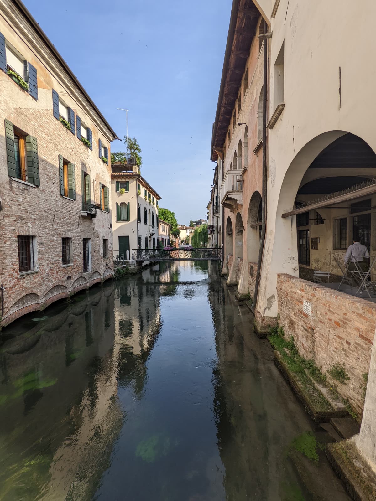 A narrow Treviso canal in the early light, still water mirroring old brick and stucco houses with green shutters and arcaded ground floors, a small iron footbridge crossing in the distance