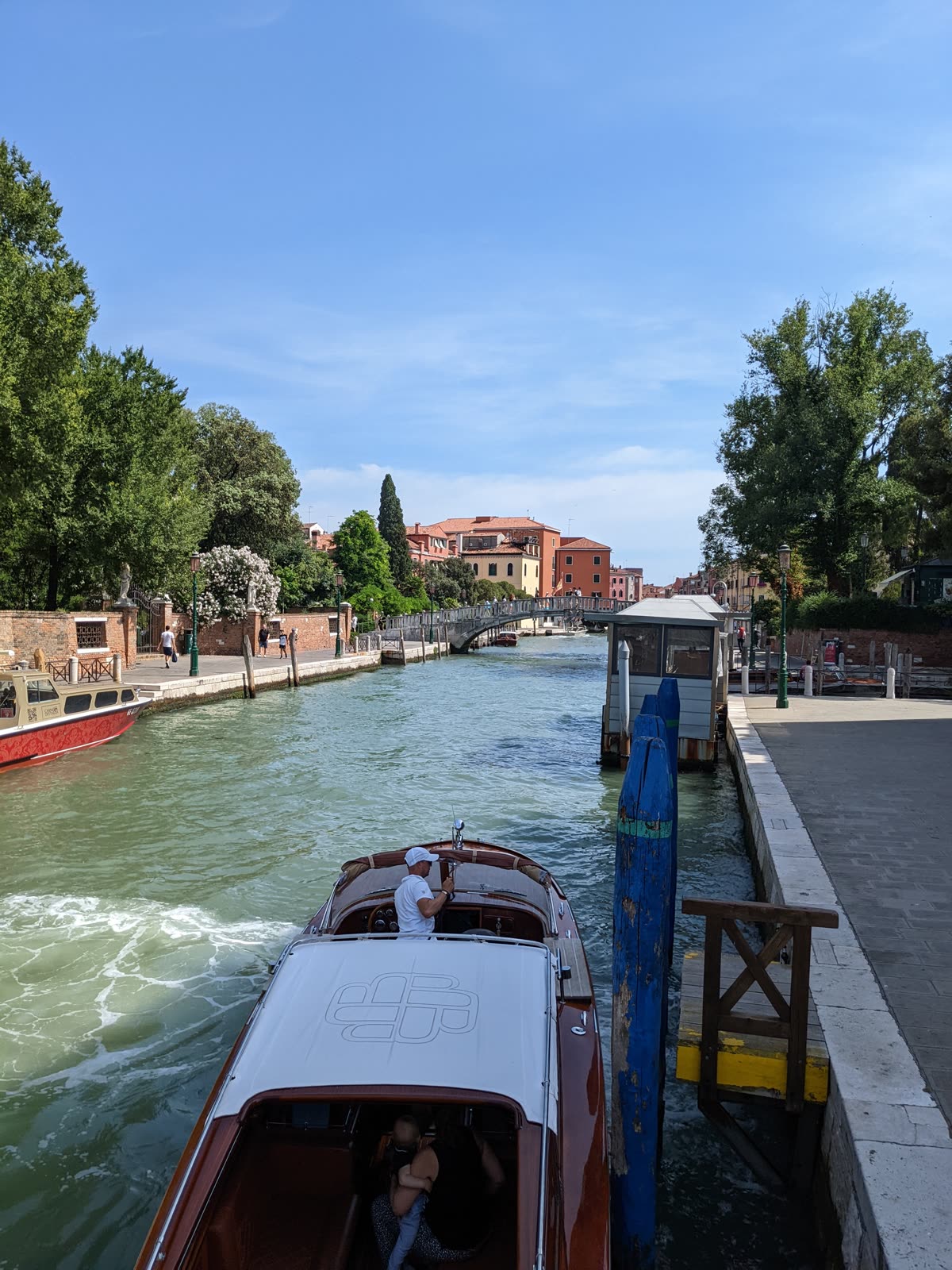 A polished wooden Venetian water-taxi pulling up to a pontoon on a side canal, its driver in white standing at the wheel, a stone footbridge and pastel palazzi ahead, trees and a garden wall along the left bank