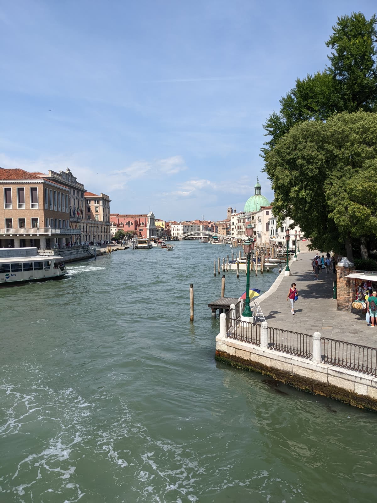 The Grand Canal in Venice under a bright blue sky, the green dome of San Simeone Piccolo on the right, the arched Ponte degli Scalzi in the distance, a vaporetto and water traffic on the busy green water, a stone fondamenta walkway with pedestrians at right