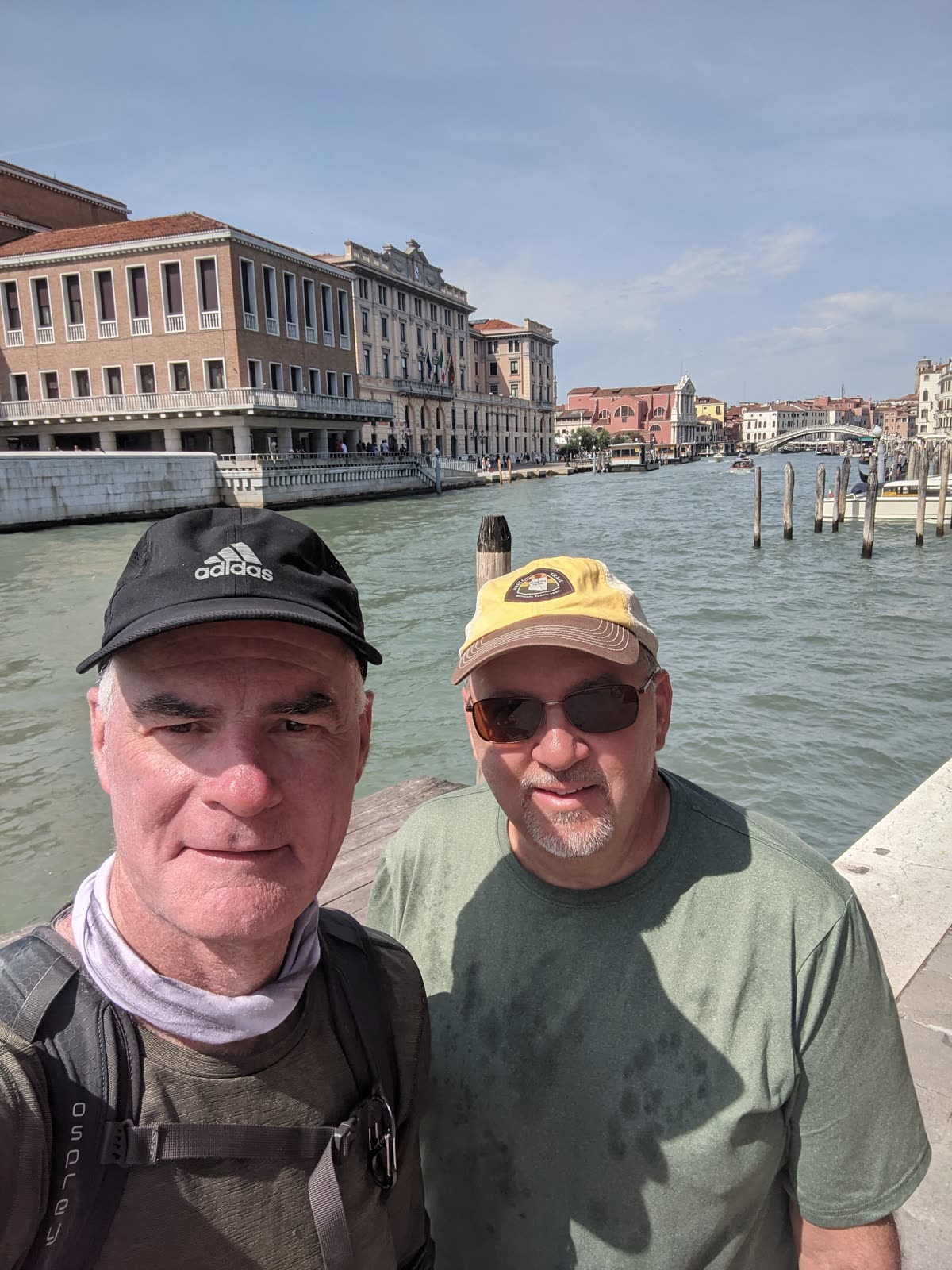 Selfie of Mike and Doug on the Grand Canal in Venice, Doug on the left in a black adidas cap with a buff at his neck and a backpack, Mike on the right in a tan cap and sunglasses with a grey goatee, the green canal, palazzi and a distant bridge behind them under blue sky