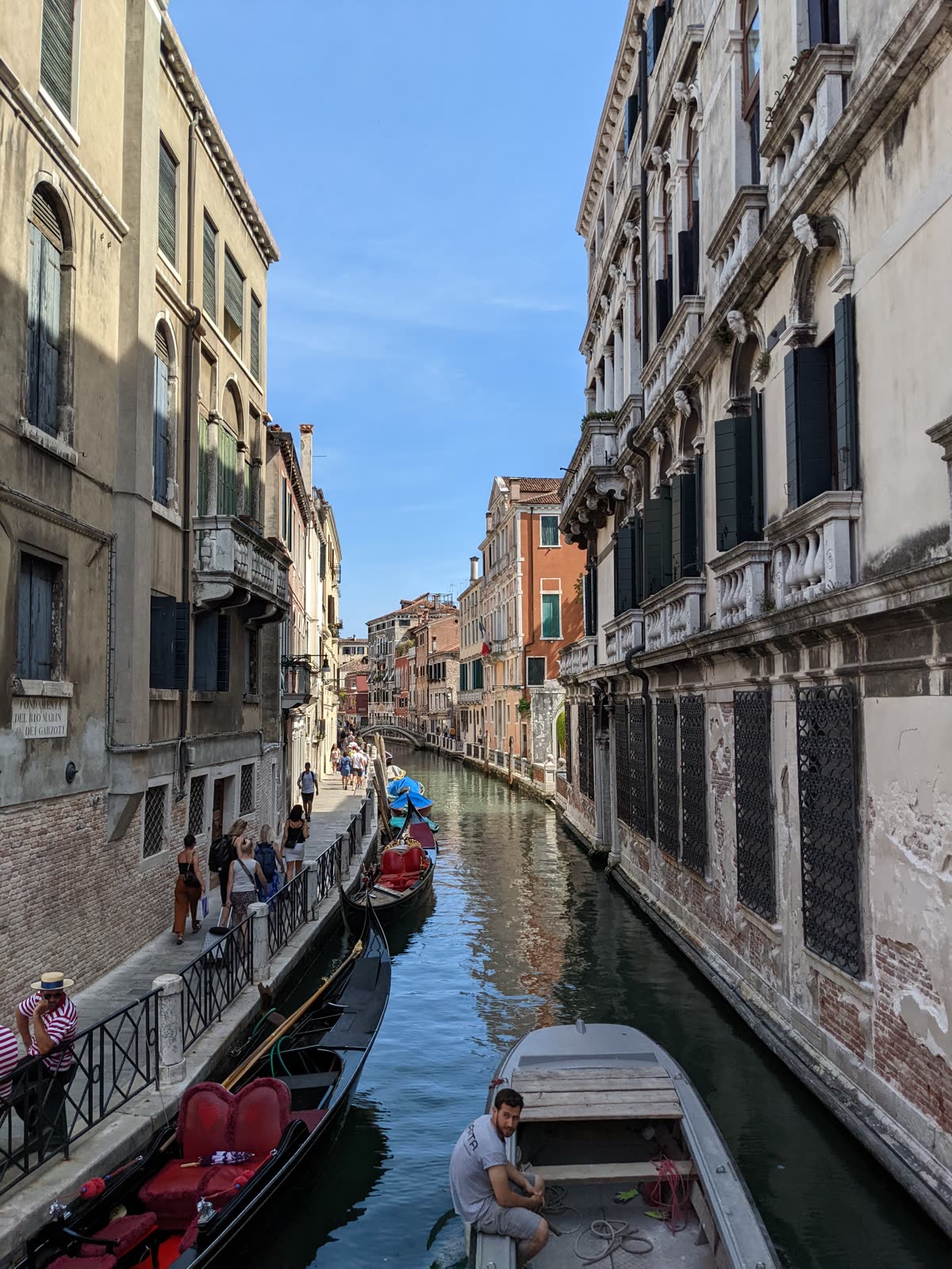 A narrow Venetian side canal, the Rio Marin, lined with moored gondolas with red velvet seats, a gondolier in a striped shirt and straw hat leaning on the railing at left, a workman in a small boat in the foreground, tall pastel palazzi and a footbridge in the distance