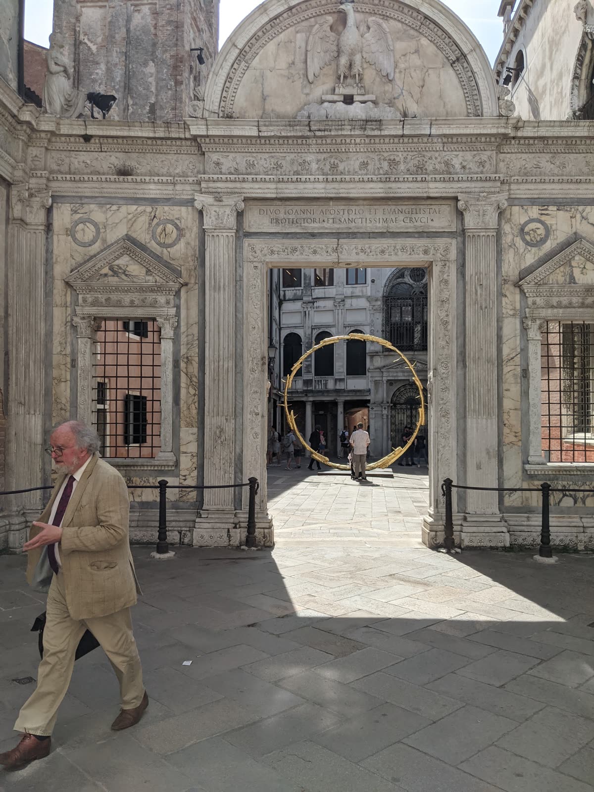 The ornate white marble screen and portal of the Scuola Grande di San Giovanni Evangelista in Venice, carved with an eagle above the arch and a Latin inscription, framing a sunlit courtyard beyond with a large gold ring sculpture, a well-dressed older man walking past in the foreground