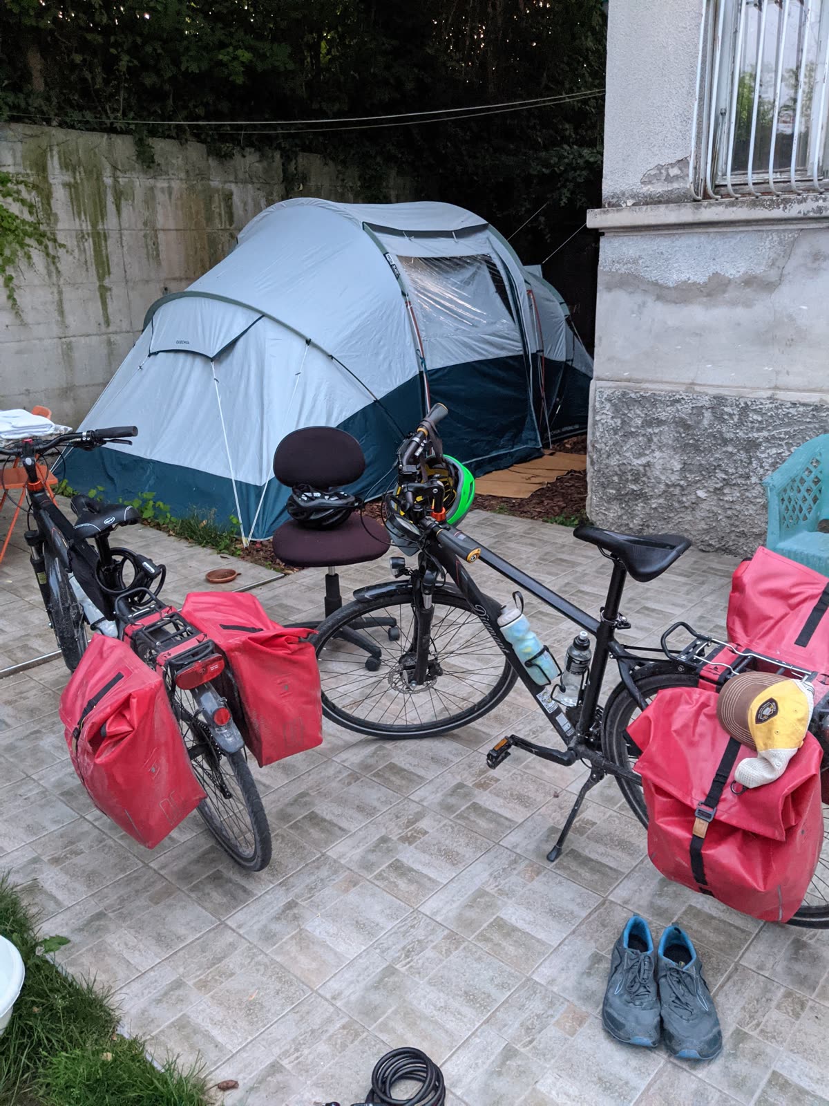 At dusk in a Verona back yard, two loaded touring bikes with red waterproof panniers and a bike helmet stand on a tiled patio, an office chair beside them, in front of a grey-and-teal dome tent pitched against a mossy concrete wall, a pair of shoes and a bike lock on the ground