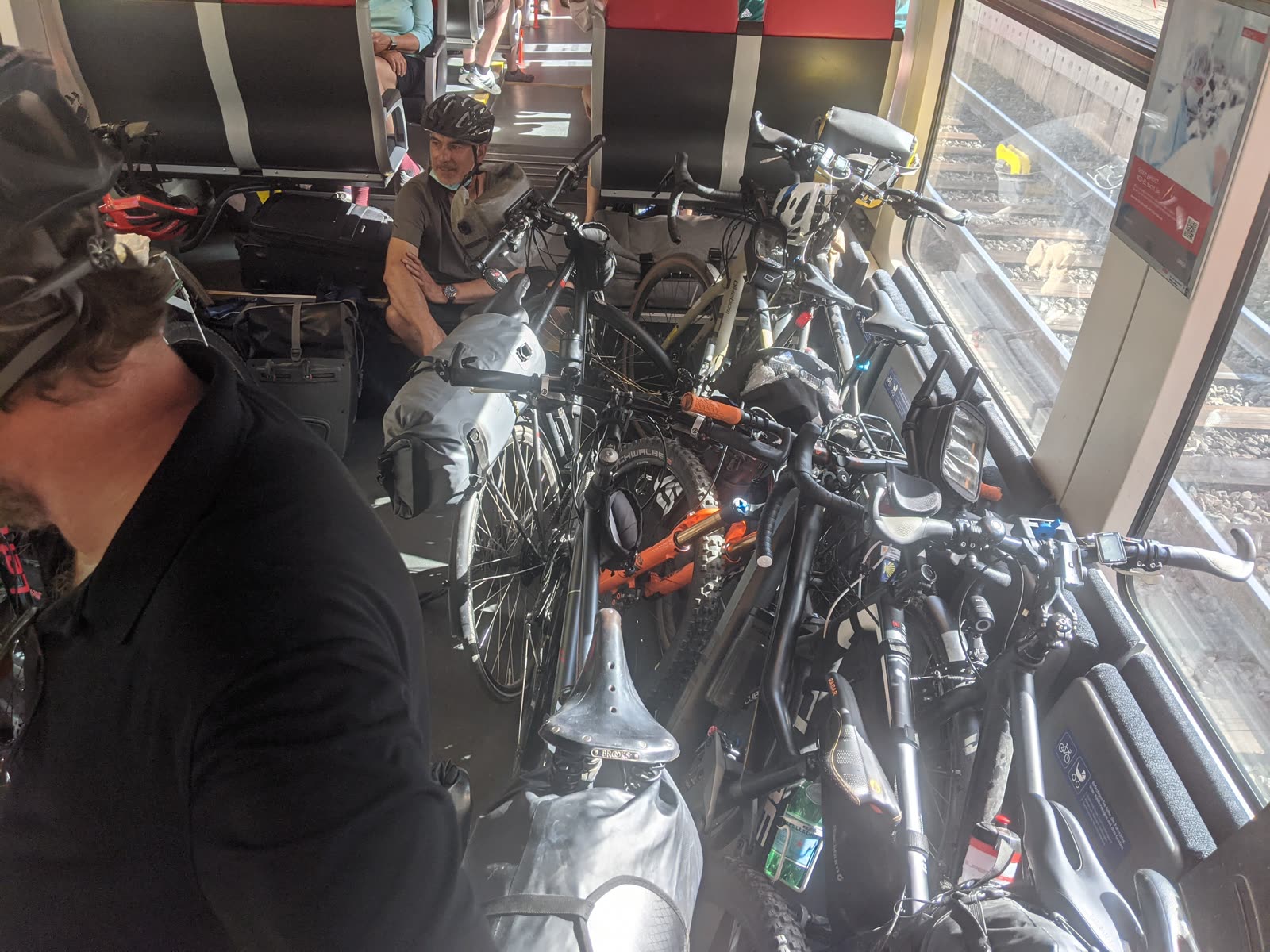The interior of a train carriage crammed wall-to-wall with loaded touring bikes and mountain bikes, panniers and dry-bags piled among them, a cyclist in a helmet squeezed into a seat, railway tracks visible through the window