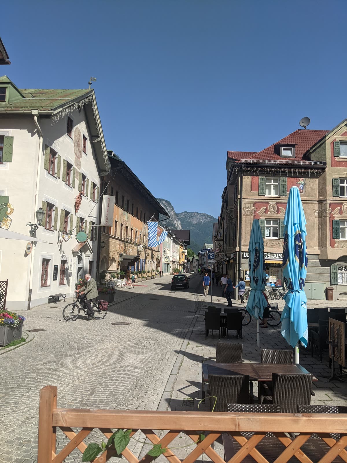Garmisch-Partenkirchen's painted main street in morning sun, Lüftlmalerei frescoes on the tall houses, blue-and-white Bavarian flags hanging out, café tables and folded umbrellas, an older man on a bicycle, forested mountains closing off the end of the street