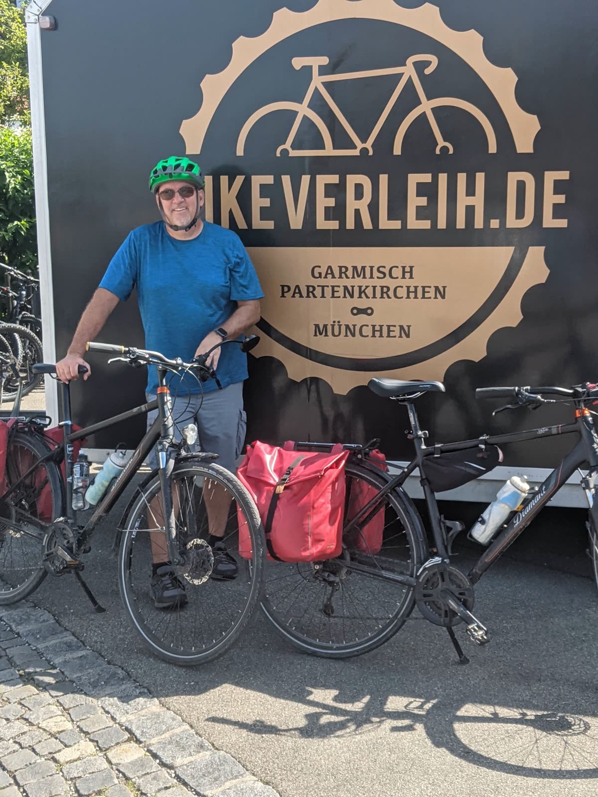 Mike in a green bike helmet and sunglasses with a grey goatee, standing and smiling behind two loaded touring bikes in front of a black trailer with a gold bikeverleih.de bicycle logo