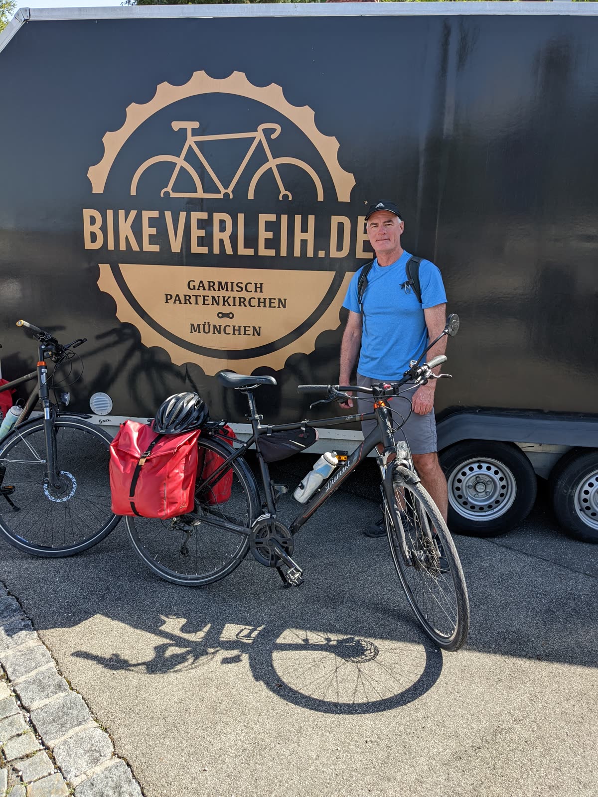 Doug in his black adidas cap and a blue shirt with a small backpack, standing beside a loaded touring bike in front of the black bikeverleih.de trailer with its gold bicycle logo