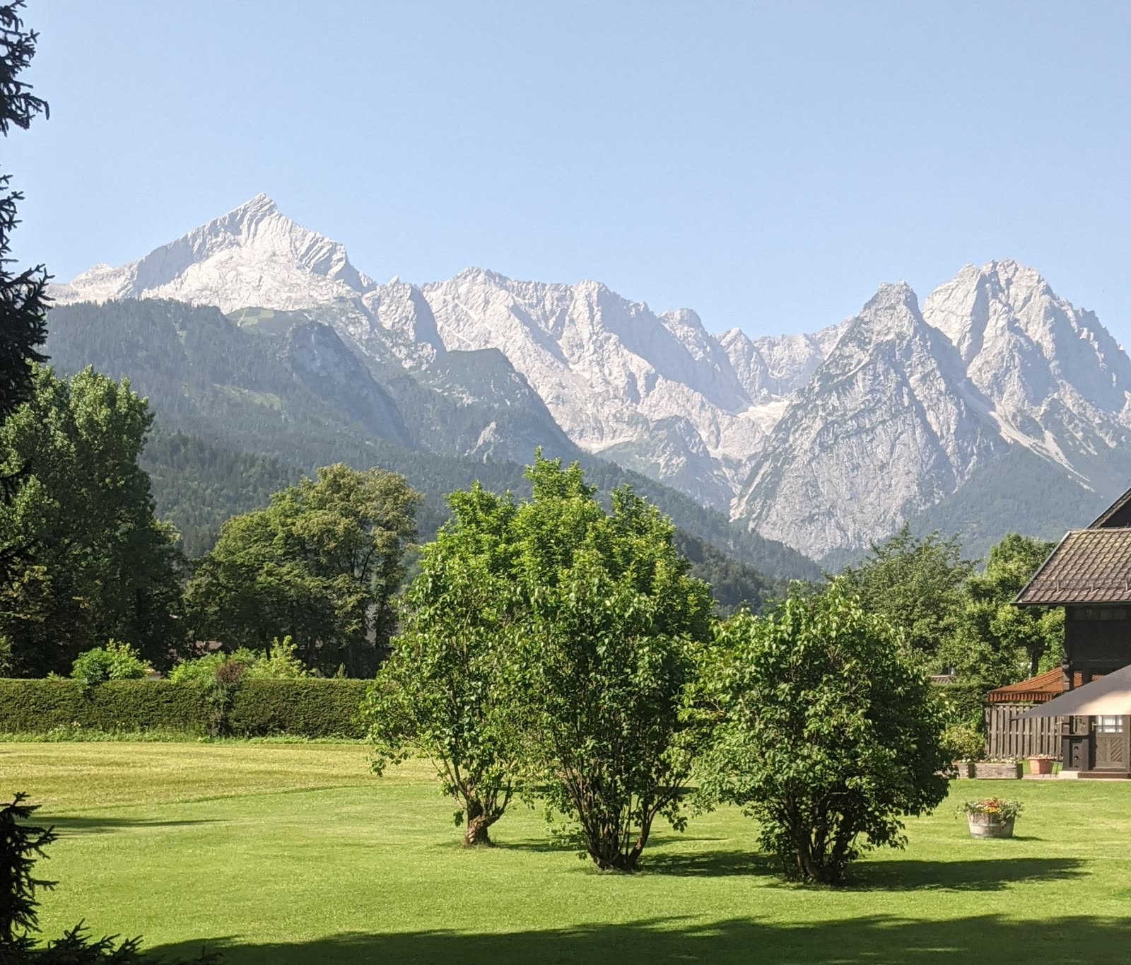 The jagged grey wall of the Wetterstein mountains, the sharp Alpspitze peak on the left, rising above forested slopes and a manicured green Garmisch lawn dotted with shrubs and trees under a clear blue sky