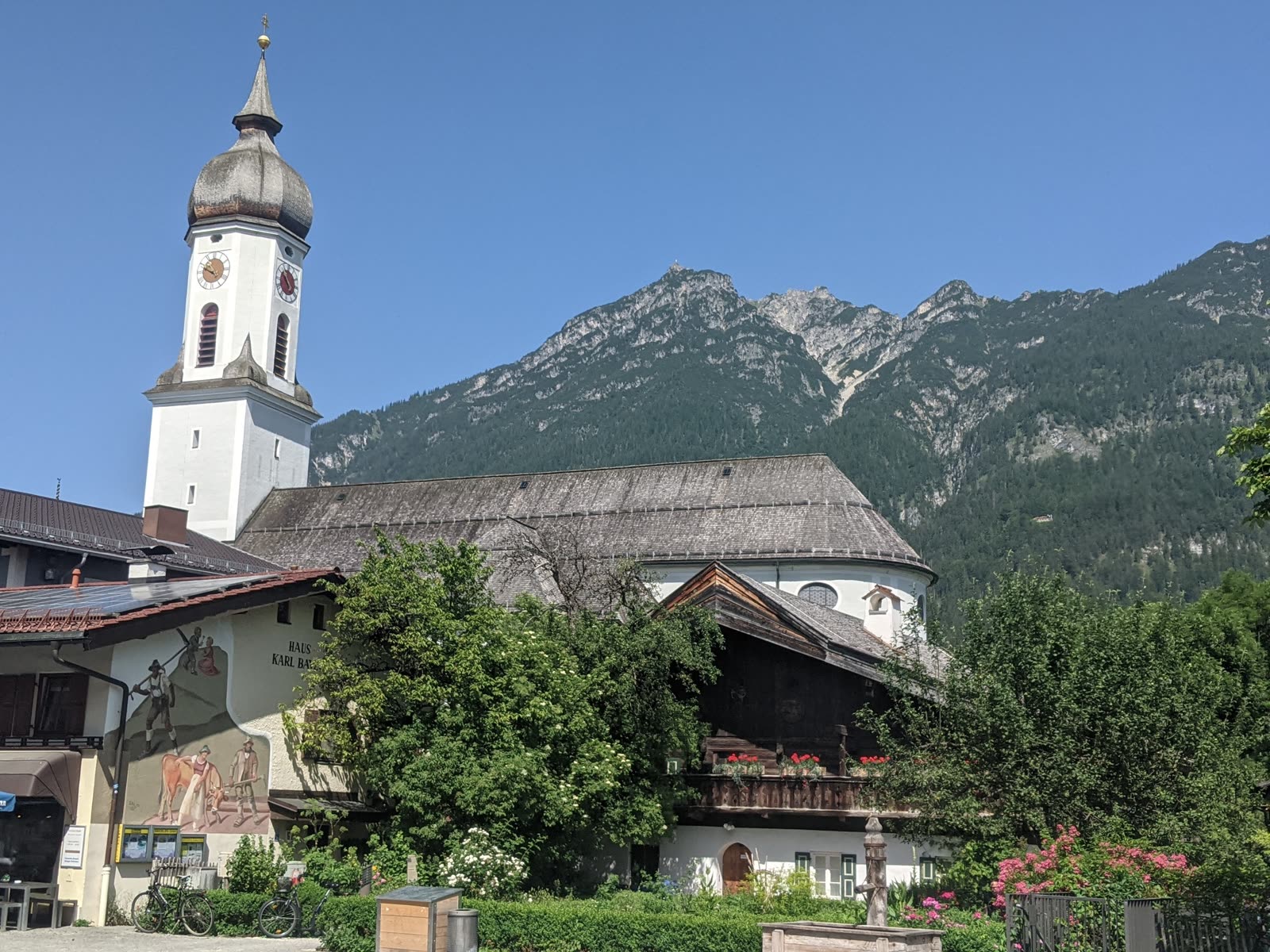 Garmisch's parish church of St. Martin with its tall white tower and grey onion dome, beside a frescoed Bavarian house, forested mountains rising behind under a clear blue sky, bicycles and flowers in the foreground
