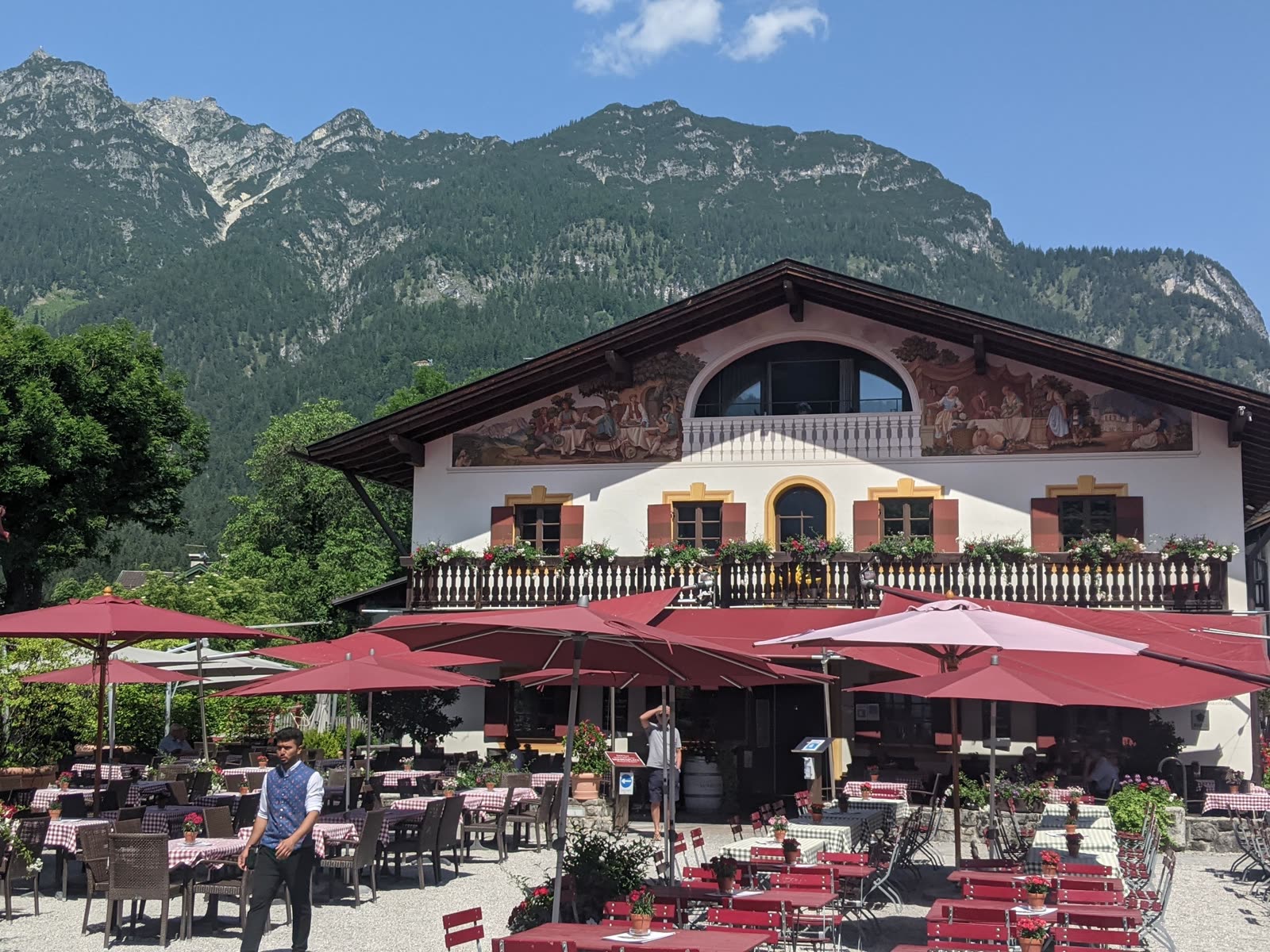 A frescoed Bavarian gasthof with a painted gable, red sun umbrellas and beer-garden tables with red-checkered cloths in front, the steep grey-green wall of the Kramer mountain rising steeply behind under blue sky