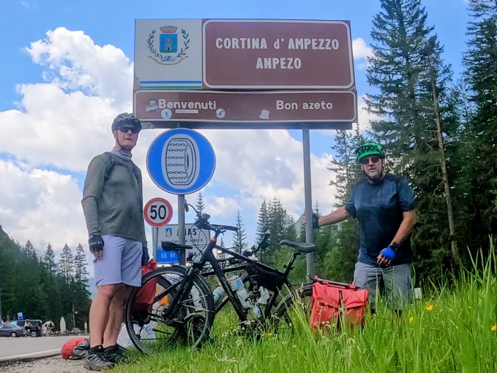 Mike and Doug standing with two loaded touring bikes beneath a brown road sign reading CORTINA d'AMPEZZO ANPEZO, forested Dolomite slopes behind