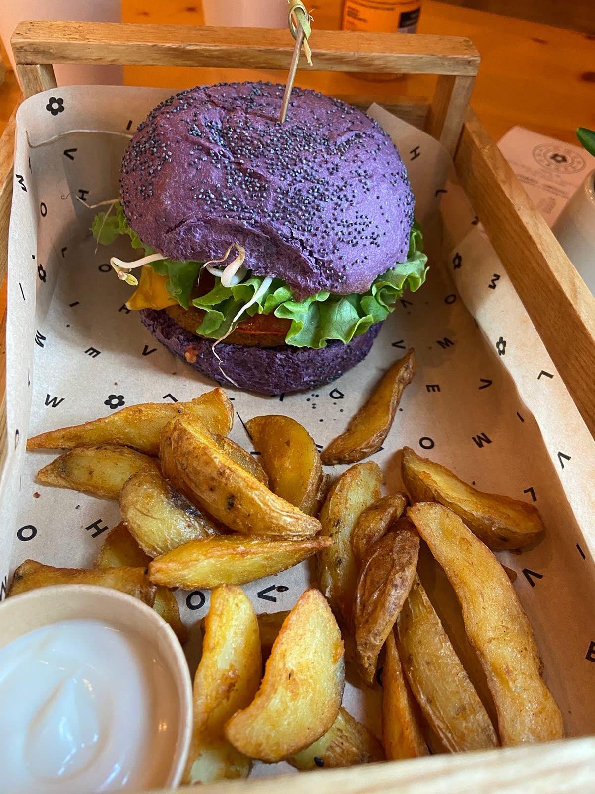 A burger on a bright purple poppy-seed bun with lettuce, sprouts, a patty and cheese, served in a wooden tray alongside golden potato wedges and a small pot of white dip