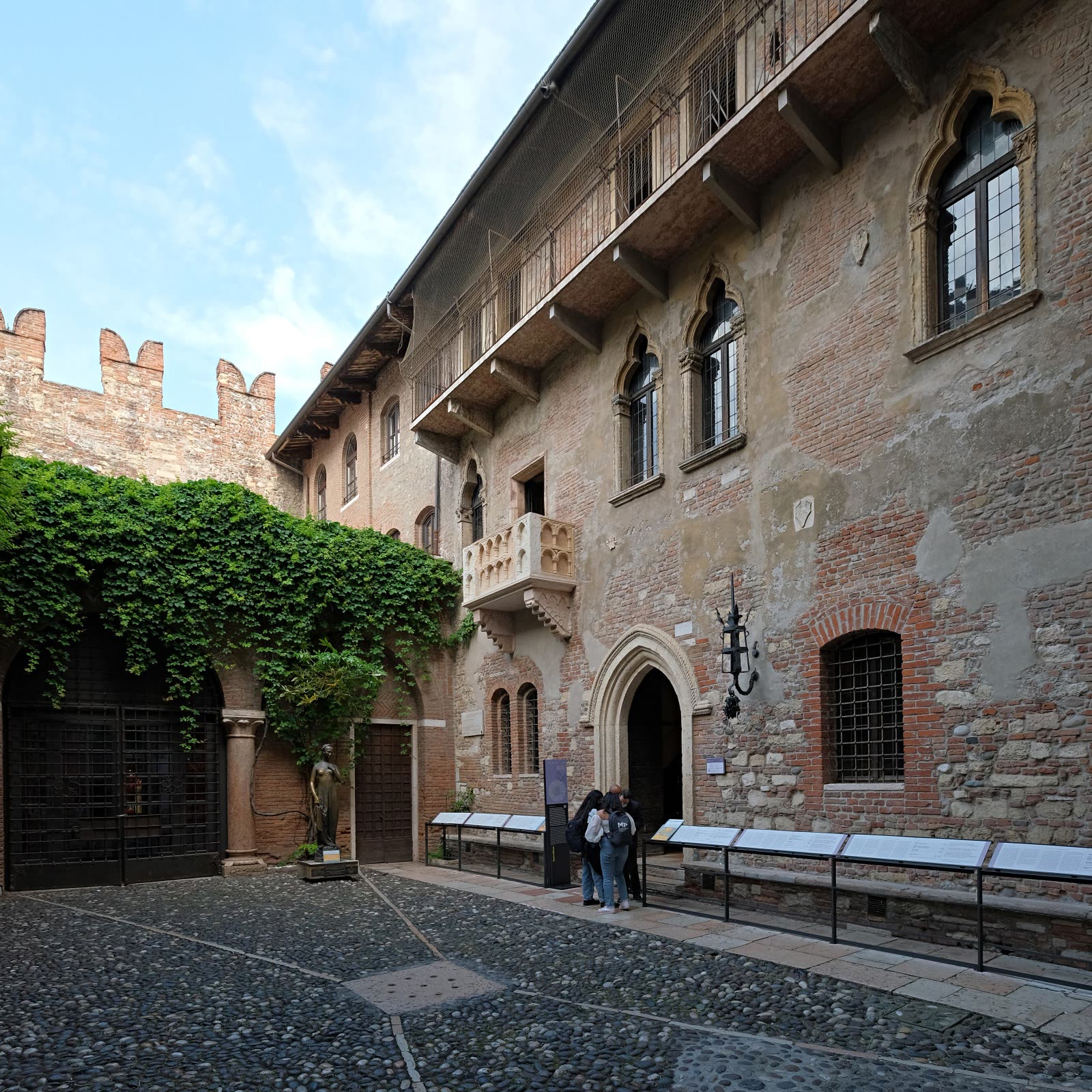The cobbled courtyard of the Casa di Giulietta in Verona, the famous small stone balcony high on a weathered brick-and-stucco wall, a bronze statue of Juliet below beside an ivy-draped archway, a few visitors reading plaques along the courtyard wall